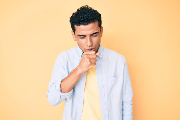 黄色の背景に立っている若いハンサムなヒスパニック系の男性は具合が悪く感じ、風邪や気管支炎の症状として咳をします。ヘルスケアの概念.  - 写真・画像
