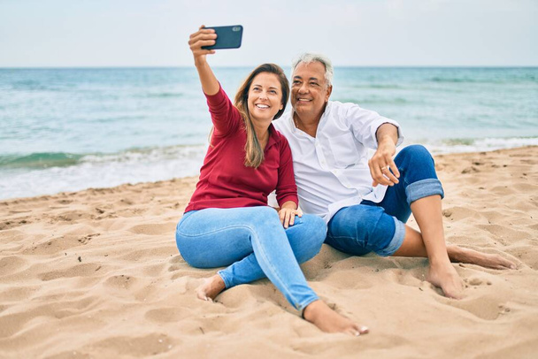 Pareja hispana de mediana edad sonriendo feliz haciendo selfie junto al smartphone en la playa. - Foto, Imagen