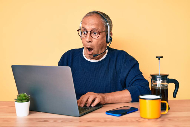 Senior handsome man with gray hair working at the office wearing operator headset scared and amazed with open mouth for surprise, disbelief face  - Foto, imagen