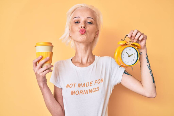 Young blonde woman with tattoo holding alarm clock and take away cup of coffee looking at the camera blowing a kiss being lovely and sexy. love expression.  - Foto, Bild