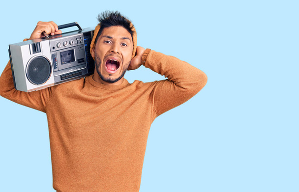 Handsome latin american young man holding boombox, listening to music crazy and scared with hands on head, afraid and surprised of shock with open mouth  - Photo, Image