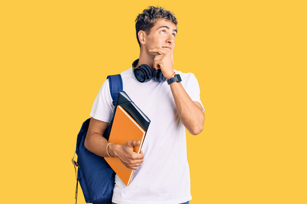 Fiatal jóképű férfi kezében diák hátizsák és könyvek komoly arc gondolkodás kérdés kézzel az állon, gondolkodó zavaros ötlet  - Fotó, kép