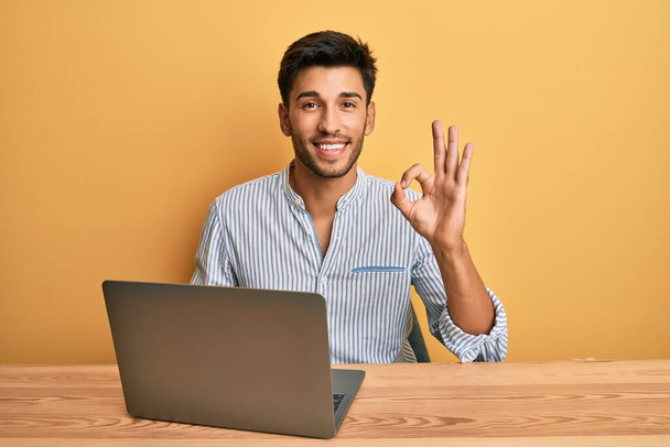 Молодий красивий чоловік, що працює в офісі з ноутбуком, робить знак ок з пальцями, посміхаючись дружній жест відмінний символ
  - Фото, зображення