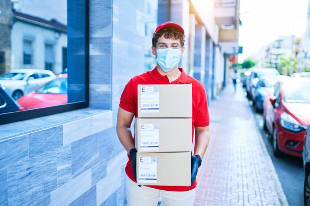 Jeune livreur caucasien portant un masque médical de protection contre le coronavirus tenant des paquets en carton marchant en ville. - Photo, image