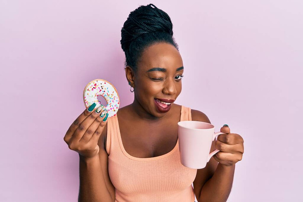 Young african american woman eating doughnut and drinking coffee winking looking at the camera with sexy expression, cheerful and happy face.  - Фото, зображення