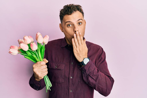 Jonge Spaanse man met boeket van roze tulpen bloemen bedekken mond met de hand, geschokt en bang voor fouten. verraste uitdrukking  - Foto, afbeelding