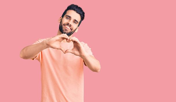 Young handsome man with beard wearing casual t-shirt smiling in love doing heart symbol shape with hands. romantic concept.  - Photo, Image