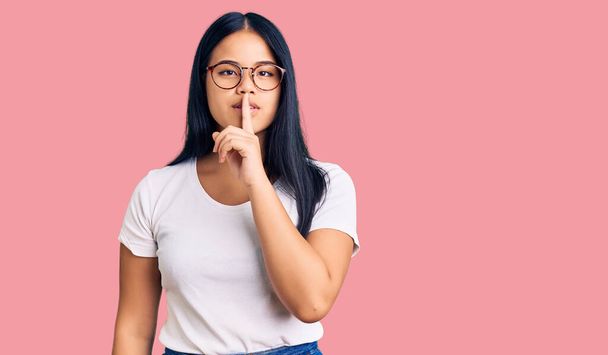 Junge schöne asiatische Mädchen tragen lässige Kleidung und Brille und bitten um Ruhe mit dem Finger auf den Lippen. Schweigen und geheimes Konzept.  - Foto, Bild