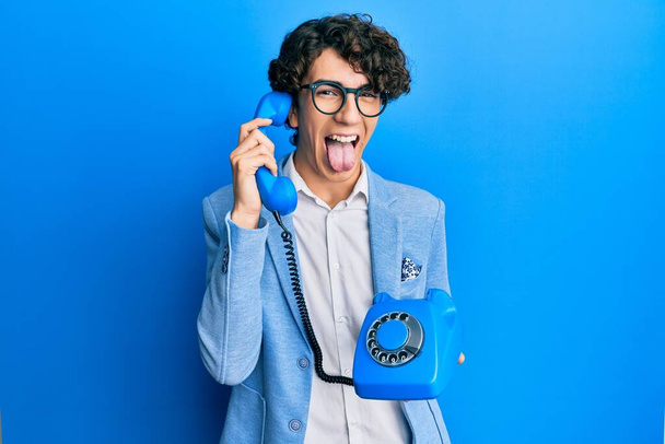 Hispanic young man speaking on vintage telephone sticking tongue out happy with funny expression.  - Foto, Imagem