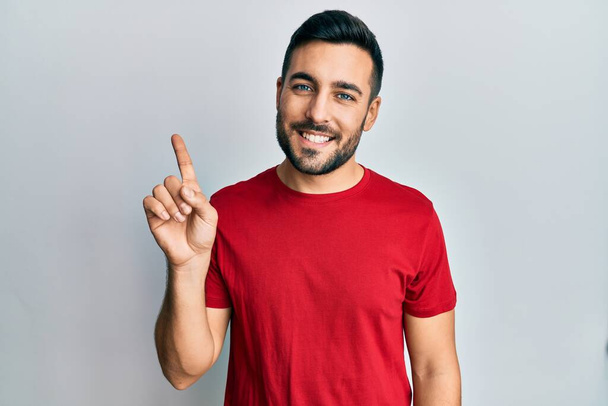 Νεαρός Ισπανός που φοράει καθημερινά ρούχα δείχνοντας και δείχνοντας με το δάχτυλο νούμερο ένα χαμογελώντας σίγουρος και χαρούμενος.  - Φωτογραφία, εικόνα