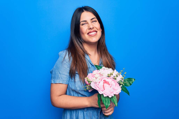 Nuori kaunis brunette nainen tilalla kimppu vaaleanpunaisia kukkia yli eristetty sininen tausta näyttää positiiviselta ja onnellinen seisova ja hymyilee luottavainen hymy osoittaa hampaat - Valokuva, kuva