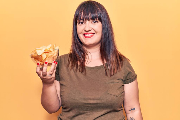 Молодая плюс размер женщина держит картофельные чипсы глядя позитивно и счастливо стоя и улыбаясь с уверенной улыбкой показывая зубы  - Фото, изображение