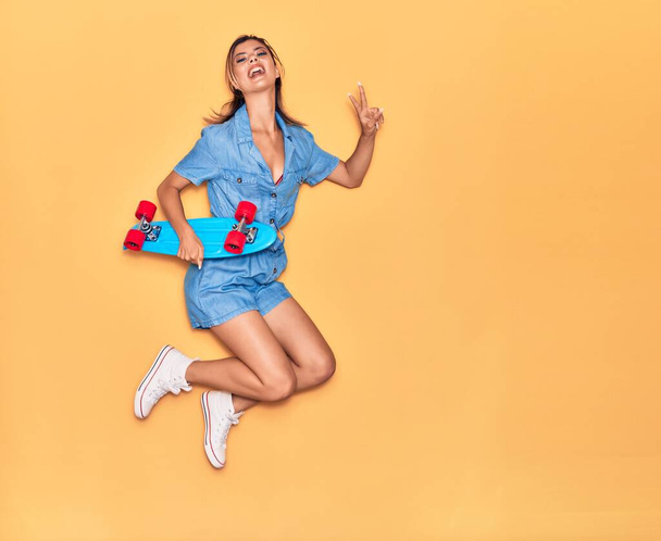Young beautiful girl holding skate smiling happy. Jumping with smile on face doing victory sign over isolated yellow background - Fotografie, Obrázek