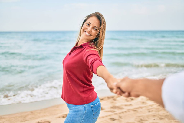 Beautiful middle age woman holding hands with husband at the beach, leading the way happy smiling in love - Фото, зображення