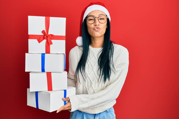 Beautiful hispanic woman wearing christmas hat and holding gifts looking at the camera blowing a kiss being lovely and sexy. love expression.  - Φωτογραφία, εικόνα