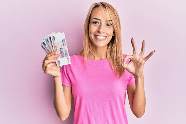 Mooie blonde vrouw met 500 Russische roebel bankbiljetten doen ok teken met de vingers, glimlachend vriendelijk gebaar uitstekend symbool  - Foto, afbeelding