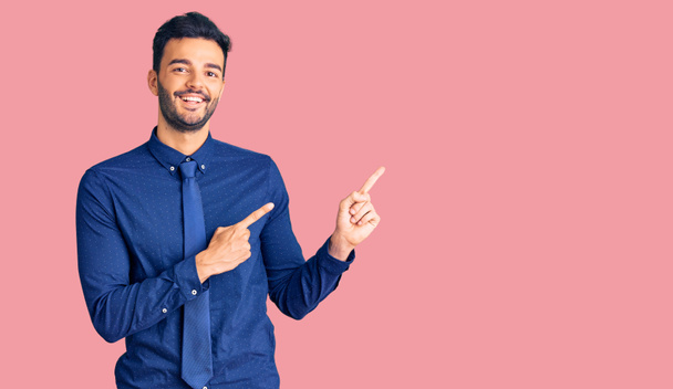 Jeune homme hispanique beau portant des vêtements de travail souriant et regardant la caméra pointant avec deux mains et doigts sur le côté.  - Photo, image