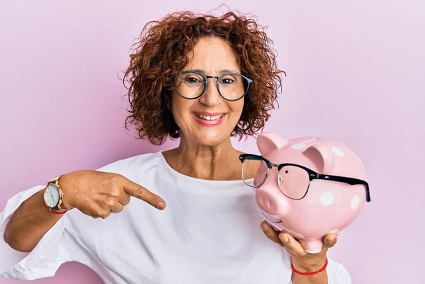 Krásná žena středního věku drží prasátko banka s brýlemi ukazující prstem na jeden self usměvavý šťastný a hrdý  - Fotografie, Obrázek