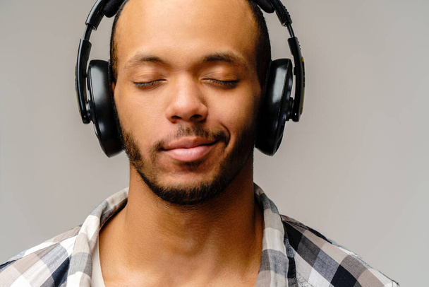 Young African-American Man Listening to Music on Large Earphones standing over light grey background - Foto, Imagem