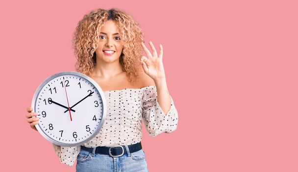 Young blonde woman with curly hair holding big clock doing ok sign with fingers, smiling friendly gesturing excellent symbol  - Фото, изображение