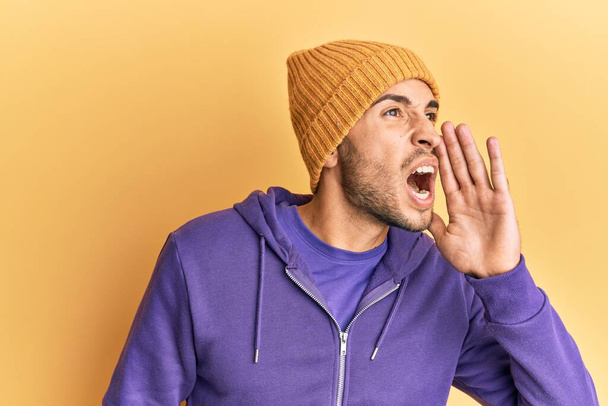 Νεαρός όμορφος άντρας που φοράει μάλλινο χειμωνιάτικο καπέλο φωνάζοντας και ουρλιάζοντας δυνατά στο πλάι με το χέρι στο στόμα. έννοια επικοινωνίας.  - Φωτογραφία, εικόνα