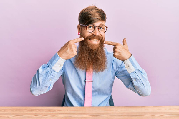 Jeune homme rousse irlandaise portant chemise d'affaires et cravate assis sur la table souriant joyeuse montrant et pointant du doigt les dents et la bouche. concept de santé dentaire.  - Photo, image