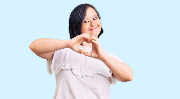 Brunette vrouw met down syndroom draagt casual witte tshirt glimlachen in liefde doen hart symbool vorm met de handen. romantisch concept.  - Foto, afbeelding