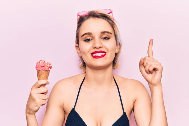 Joven hermosa mujer rubia con bikini sosteniendo helado sorprendido con una idea o pregunta apuntando con el dedo con la cara feliz, número uno  - Foto, imagen