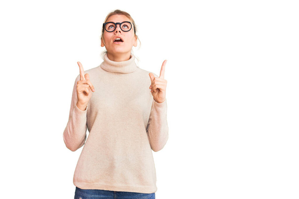 Young beautiful blonde woman wearing turtleneck sweater and glasses amazed and surprised looking up and pointing with fingers and raised arms.  - Foto, Imagen