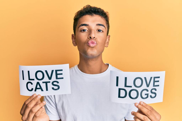 Νεαρός όμορφος Αφροαμερικάνος που κρατάει χαρτί μαζί του αγαπώ τις γάτες και λατρεύω τις σκυλίσιες φράσεις κοιτάζοντας την κάμερα να φυσάει ένα φιλί όντας αξιαγάπητος και σέξι. έκφραση αγάπης.  - Φωτογραφία, εικόνα