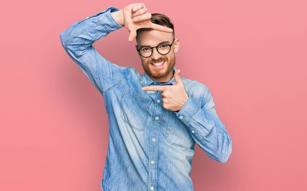 Молодой рыжий мужчина в джинсовой рубашке, улыбающийся, делая рамку с руками и пальцами со счастливым лицом. креативность и концепция фотографии.  - Фото, изображение