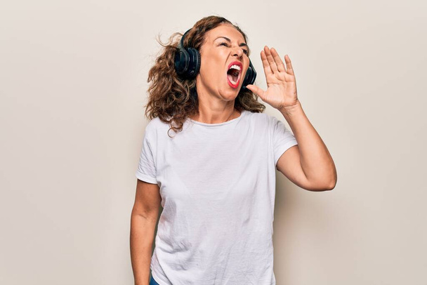 Moyen Age belle femme écoutant de la musique à l'aide d'écouteurs sur fond blanc criant et criant fort à côté avec la main sur la bouche. Concept de communication. - Photo, image