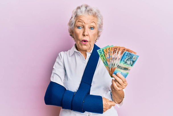 Donna anziana dai capelli grigi che indossa un braccio sulla fionda con banconote in dollari australiani in faccia shock, dall'aspetto scettico e sarcastico, sorpresa a bocca aperta  - Foto, immagini