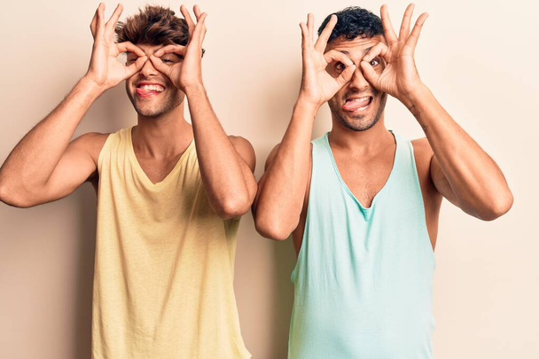 Young gay couple wearing casual clothes doing ok gesture like binoculars sticking tongue out, eyes looking through fingers. crazy expression.  - Фото, зображення
