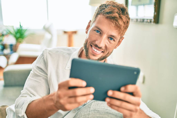 Красивий білий чоловік посміхається щасливо сидячи на дивані вдома, використовуючи пристрій тачпад
 - Фото, зображення