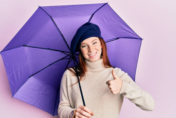 Genç kızıl saçlı kız elinde mor şemsiye, yeni bere takmış mutlu ve pozitif gülümsüyor, başparmağıyla mükemmel ve onay işareti yapıyor.  - Fotoğraf, Görsel