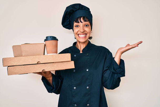 Όμορφη brunette γυναίκα επαγγελματίας σεφ κρατώντας πάρει μακριά το φαγητό γιορτάζει τη νίκη με χαρούμενο χαμόγελο και την έκφραση νικητής με υψωμένα χέρια  - Φωτογραφία, εικόνα
