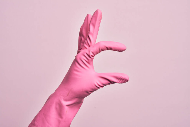 Ręka białego młodzieńca z rękawicą czyszczącą nad odizolowanym różowym tłem zbierającym i zabierającym niewidzialną rzecz, trzymającym obiekt z palcami pokazującymi przestrzeń - Zdjęcie, obraz