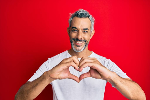 Hombre de pelo gris de mediana edad vistiendo una camiseta blanca casual sonriendo enamorado mostrando el símbolo del corazón y la forma con las manos. concepto romántico.  - Foto, Imagen