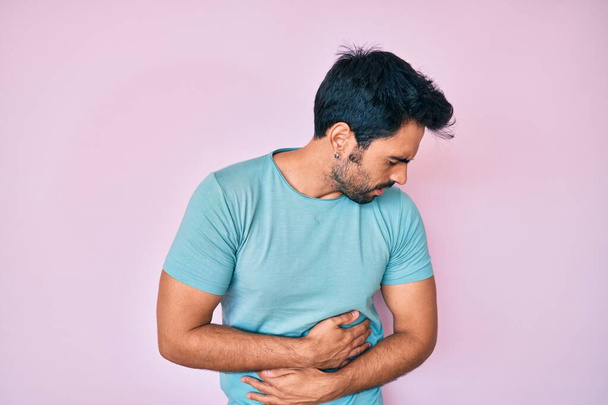 Handsome hispanic man with beard wearing casual clothes with hand on stomach because indigestion, painful illness feeling unwell. ache concept.  - Foto, Bild