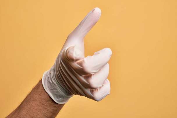 İzole edilmiş sarı arka plan üzerinde tıbbi eldiveni olan beyaz bir gencin eli kameraya işaret ediyor ve yönü gösteriyor. - Fotoğraf, Görsel