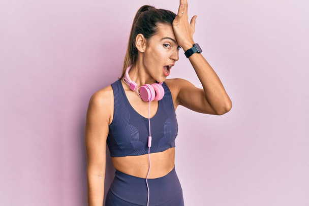 Young beautiful woman wearing gym clothes and using headphones surprised with hand on head for mistake, remember error. forgot, bad memory concept.  - Photo, Image