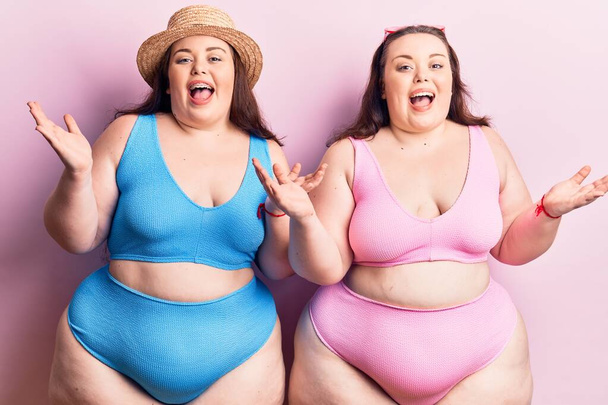 Jóvenes gemelos más tamaño vistiendo bikini celebrando la victoria con sonrisa feliz y expresión ganadora con las manos levantadas  - Foto, Imagen
