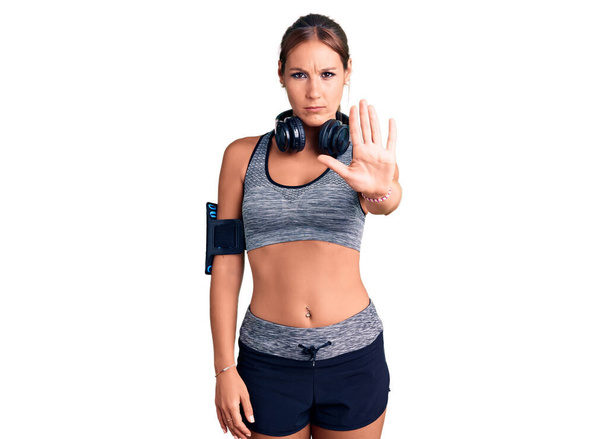 Young beautiful hispanic woman wearing gym clothes and using headphones doing stop sing with palm of the hand. warning expression with negative and serious gesture on the face.  - Photo, Image