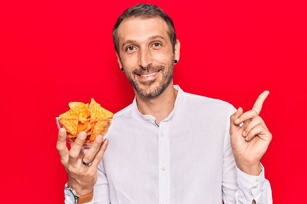 Junger gutaussehender Mann mit Nachos Kartoffelchips in der Hand lächelt glücklich und zeigt mit der Hand und dem Finger zur Seite  - Foto, Bild