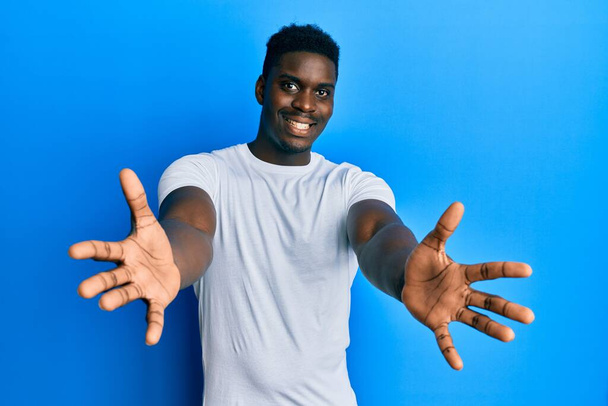 Handsome black man wearing casual white t shirt looking at the camera smiling with open arms for hug. cheerful expression embracing happiness.  - Фото, изображение