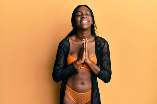 Jovem afro-americana vestindo lingerie implorando e orando com as mãos em conjunto com a expressão da esperança no rosto muito emocional e preocupado. implorando.  - Foto, Imagem