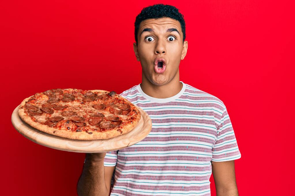 Fiatal arab férfi kezében olasz pizza ijedt és csodálkozott nyitott szájjal meglepetés, hitetlenség arc  - Fotó, kép