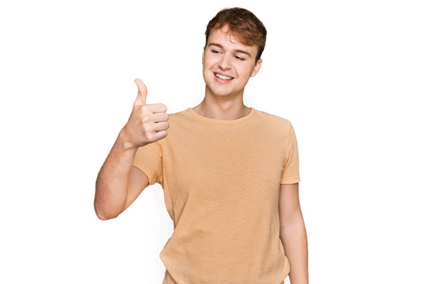 Jovem caucasiano vestindo roupas casuais que parecem orgulhosos, sorrindo fazendo polegares até gesto para o lado  - Foto, Imagem