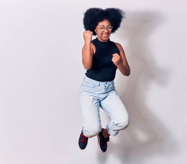 Young beautiful african american woman wearing casual clothes and glasses smiling happy. Jumping with smile on face doing winner gesture with fists up over isolated white background - 写真・画像
